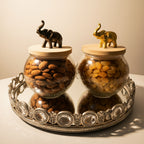 Two glass jars with wooden lids containing almonds and raisins, each topped with an elephant figurine, on a decorative tray.